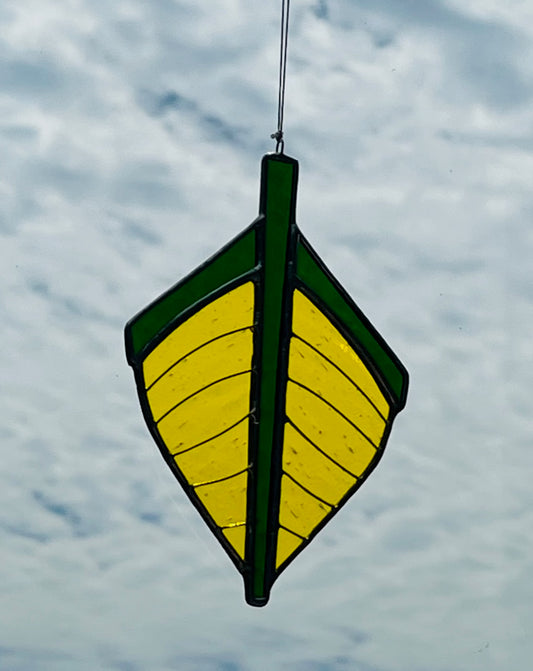 Our traditional punt-shaped stained glass suncatcher is yellow with a dark green trim. Hung with a blue sky background.