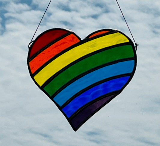 A heart shape suncatcher made with rainbow color stripes hung with a blue sky background.