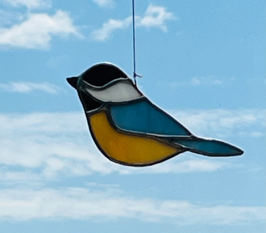 A stained glass black capped chickadee with a white neck, light blue back and tan underbelly hung with a blue sky background.
