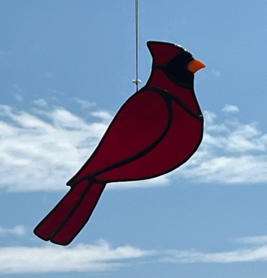 A stained glass cardinal. It is red with a black face and orange beak hung against a blue sky with white clouds background.