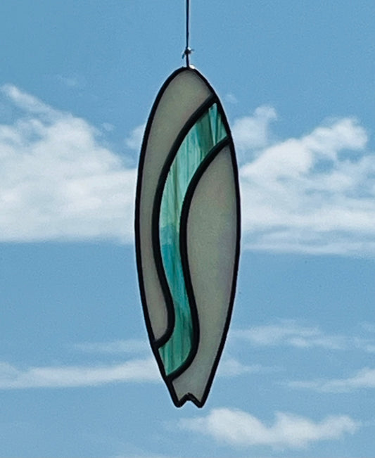 A stained glass shortboard that is made with white opalescent glass with a teal glass wave down the center. Hung with a blue sky background.