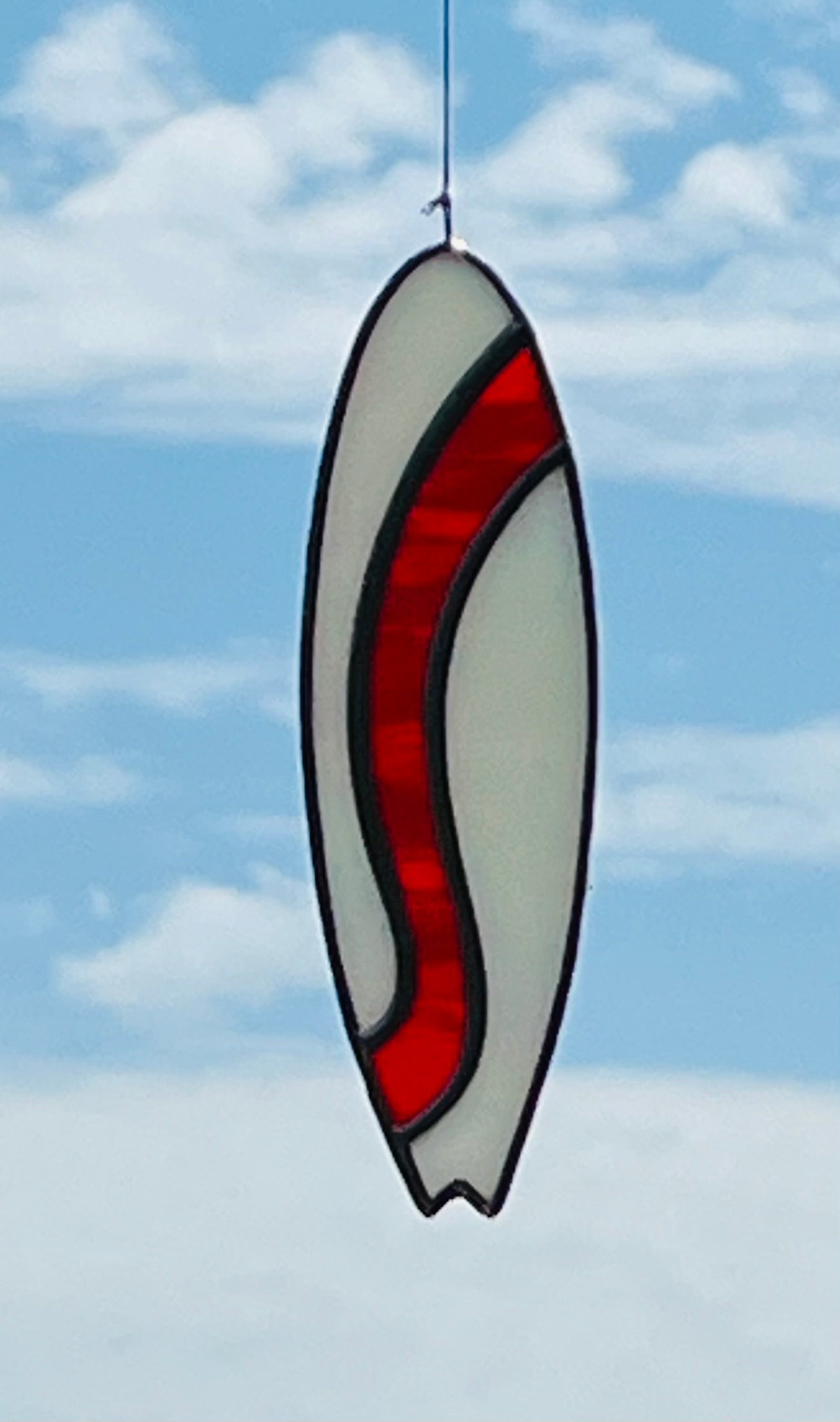 A stained glass shortboard that is made with white opalescent glass with a red glass wave down the center. Hung with a blue sky background.