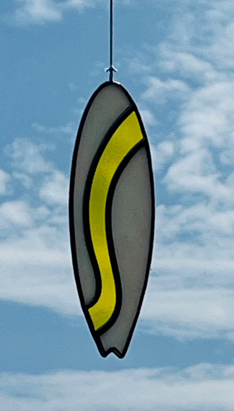 A stained glass shortboard that is made with white opalescent glass with a yellow glass wave down the center. Hung with a blue sky background.