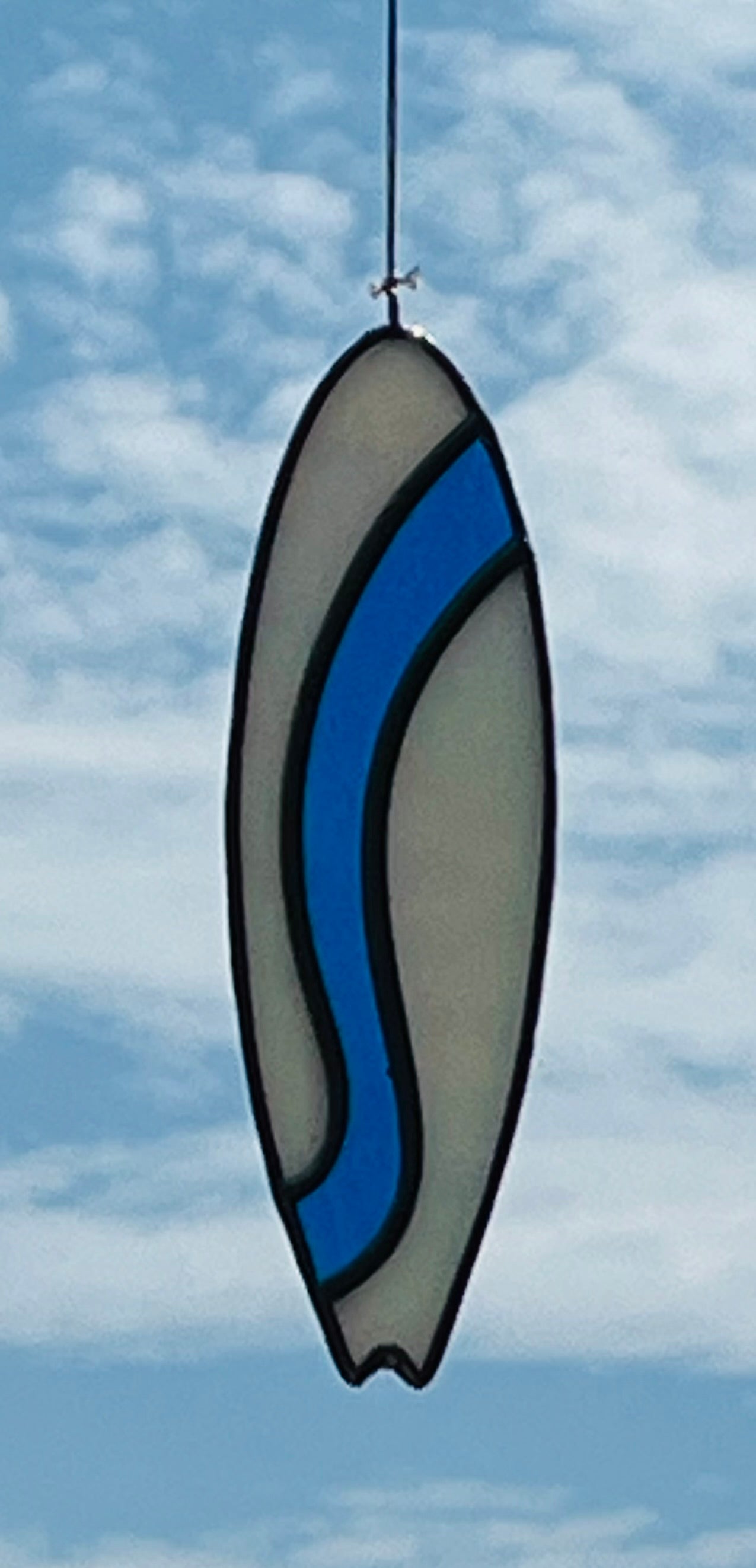 A stained glass shortboard that is made with white opalescent glass with a blue glass wave down the center. Hung with a blue sky background.