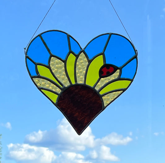 A yellow sunflower with a ladybug sitting on a petal and blue background inside a heart shaped suncatcher. Hanging Infront of blue sky and fluffy white clouds.